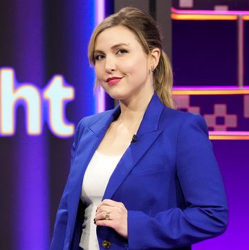 taylor tomlinson stands in front of a colorful background and smiles as she looks at the camera, she wears a royal blue suit jacket over a white top and holds one side of her jacket