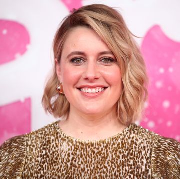 greta gerwig smiles at the camera, she wears a gold and white dress with gold hoop earrings, her shoulder length hair is styled down and she has on makeup
