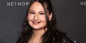 gypsy rose blanchard smiles at the camera while standing in front of a black background with white writing, she wears a black vneck dress and a necklace