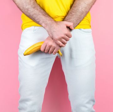 potency and men's health a man in white jeans, legs apart, holds a banana near the genitals pink background close up of hands