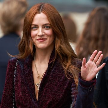 riley keough smiles and waves at the camera, she wears a burgundy jacket and golden star necklace