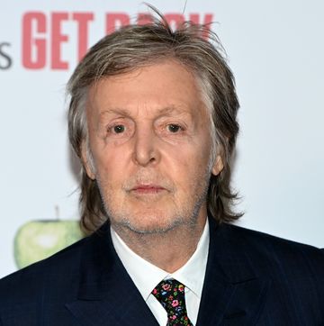 paul mccartney looks into the camera, he wears a blue suit jacket, white collared shirt and multicolored floral tie