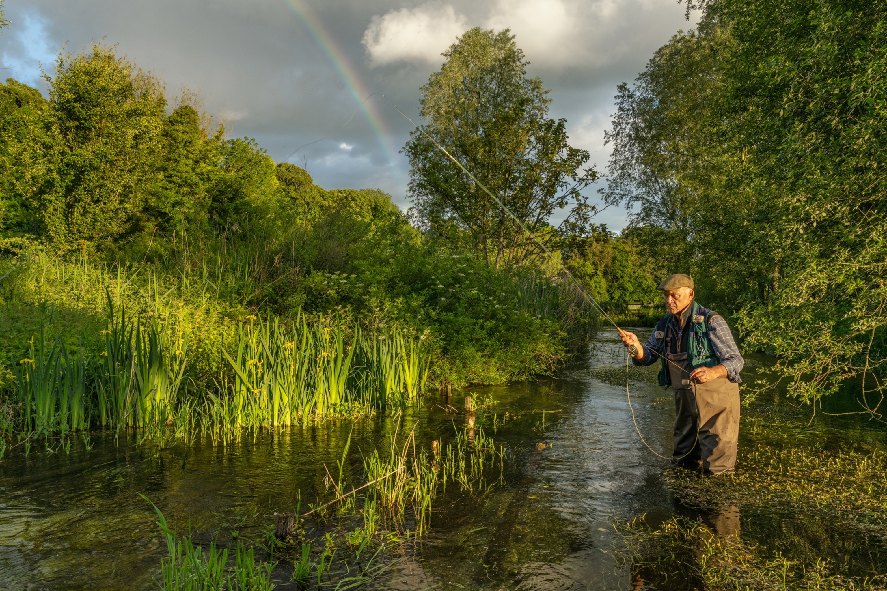 Simon is knee-deep fishing in the stream. A rainbow is behind him in the top left corner of the frame.