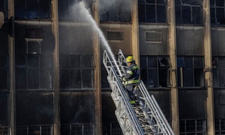 A firefighter standing on an engine ladder directs a hose at the building