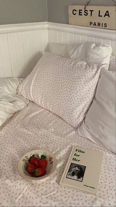 a bowl of strawberries sits on a bed with white sheets and pillows next to a book