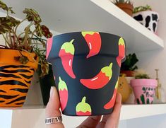 a hand holding up a cup with peppers on it in front of some potted plants