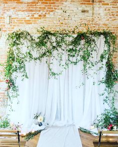 an outdoor ceremony with white drapes and greenery on the wall, along with wooden benches