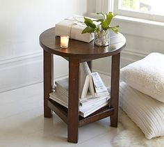 a small table with some books and a candle on it next to a window in a white room
