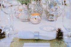 the table is set with silver and white dishes, wine glasses, napkins, and pine cones