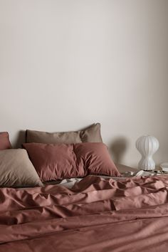 an unmade bed with red sheets and pillows in a white walled room next to a lamp