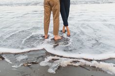 two people standing in the water holding hands and looking at the ocean with waves crashing on them