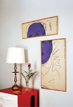 two paintings hang on the wall next to a red table with a lamp and vase