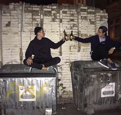 two people sitting on top of trash cans holding beer bottles in their hands and one person reaching for the bottle