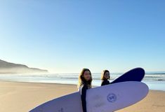two women are standing on the beach with their surfboards