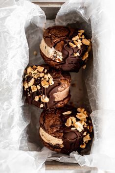 three chocolate covered donuts with nuts on top in a paper wrapper lined box