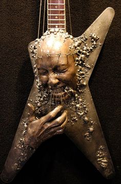 a metal sculpture of a man's face with his hands resting on the neck of an electric guitar