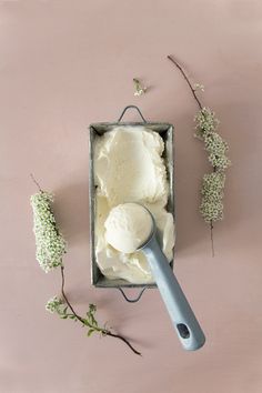 a scoop of ice cream in a tin next to flowers on a pink surface with a blue spoon