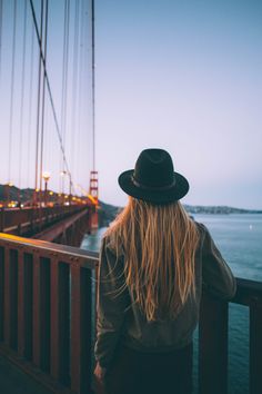 a woman with long hair wearing a hat looking at the golden gate bridge