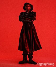 a woman in a black coat and boots standing against a red background with her arms crossed