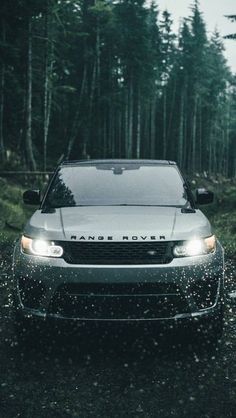 the front end of a silver car driving on a wet road near some trees and bushes