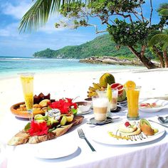 the table is set up on the beach for an outdoor meal with fruit and drinks