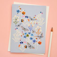 a card with butterflies and flowers on it next to a pencil, which is laying on a pink surface