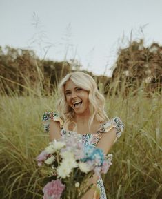 a woman standing in tall grass holding a bunch of flowers and smiling at the camera