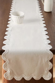 a white table runner with scalloped edges on a wooden dining room table in front of a vase and pitcher