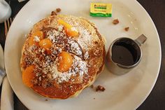 a white plate topped with pancakes covered in powdered sugar and orange slices next to a cup of coffee