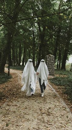 two people dressed in ghost costumes walking down a path through the woods with trees behind them