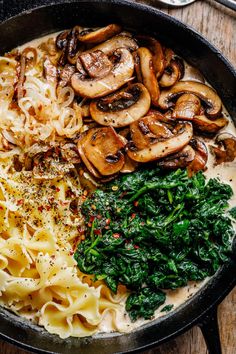 a pan filled with pasta, spinach and mushrooms on top of a wooden table