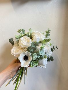 a person holding a bouquet of white flowers and greenery in their hand against a wall