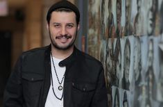 a man wearing a black jacket standing next to a wall with graffiti on it and smiling