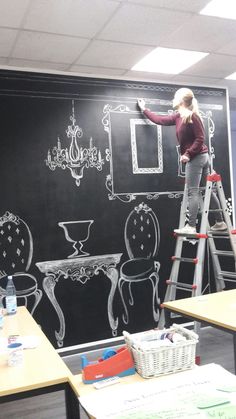 a woman standing on a ladder in front of a chalkboard with chairs and tables