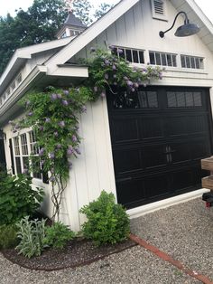 a white house with purple flowers growing on it's side and a black garage door
