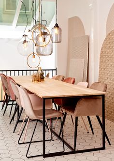 a wooden table surrounded by pink chairs and hanging light fixtures in a room with white tile flooring
