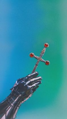 a hand that is holding a cross with red stones on the top and bottom, in front of a blue sky
