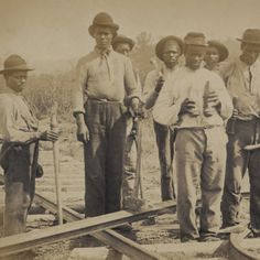 a group of men standing next to each other in front of train tracks and holding shovels