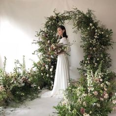 a woman standing in front of a floral arch with flowers on it and greenery around her