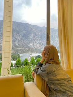 a woman sitting on top of a yellow couch in front of a window next to a lush green hillside