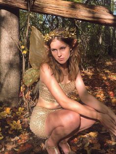 a woman dressed as a fairy sitting on the ground next to a tree with leaves around her