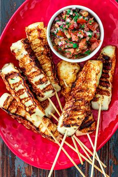 several skewered meats on a red plate with a small bowl of salsa