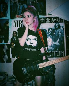 a girl with pink hair is holding a guitar and posing for the camera in front of posters