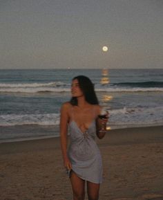 a woman standing on top of a beach next to the ocean holding a glass of wine