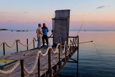 two people are standing on a dock by the water at sunset or dawn, looking out to sea