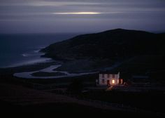 a house sitting on top of a hill next to the ocean at night with an overcast sky