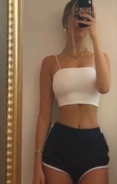 a woman taking a selfie in front of a mirror wearing black shorts and a white crop top