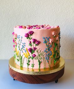 a pink and gold decorated cake on a wooden stand next to a white wall with flowers painted on it