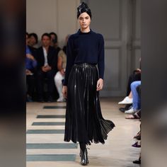 a model walks down the runway in a pleated skirt and turtle neck sweater, while people watch