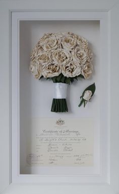 a bouquet of white roses is in a frame with a certificate on the wall behind it
